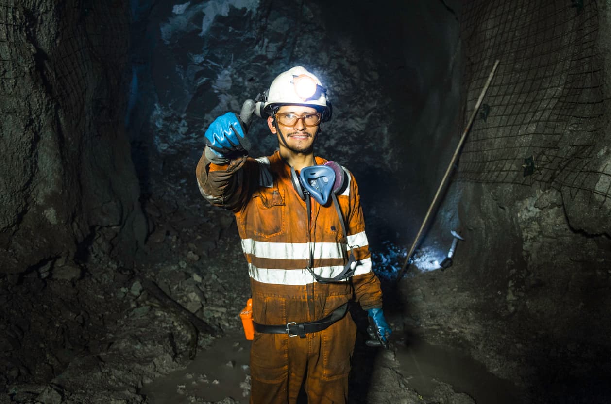 Un trabajador de minería viste un overol naranja con franjas reflectantes y se encuentra en una caverna oscura. Lleva casco con luz, gafas de seguridad y una mascarilla. Sonríe y hace un gesto de pulgar hacia arriba. En el fondo se pueden ver paredes rocosas y equipo de minería.