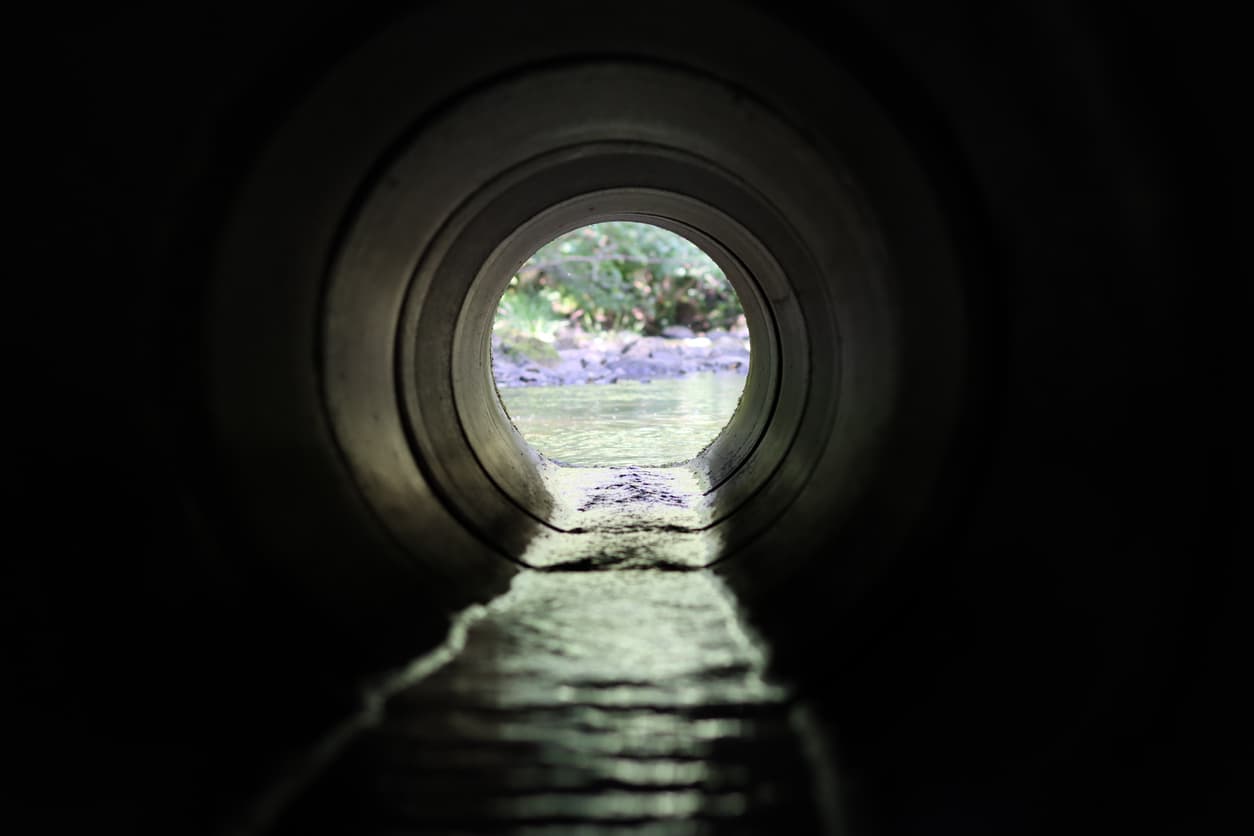 Se observa una tubería helicoidal desde el interior, con vista hacia el exterior, donde se aprecia agua fluyendo y vegetación al fondo. La luz ilumina parcialmente el interior de la tubería, creando un contraste entre la oscuridad y el paisaje brillante.