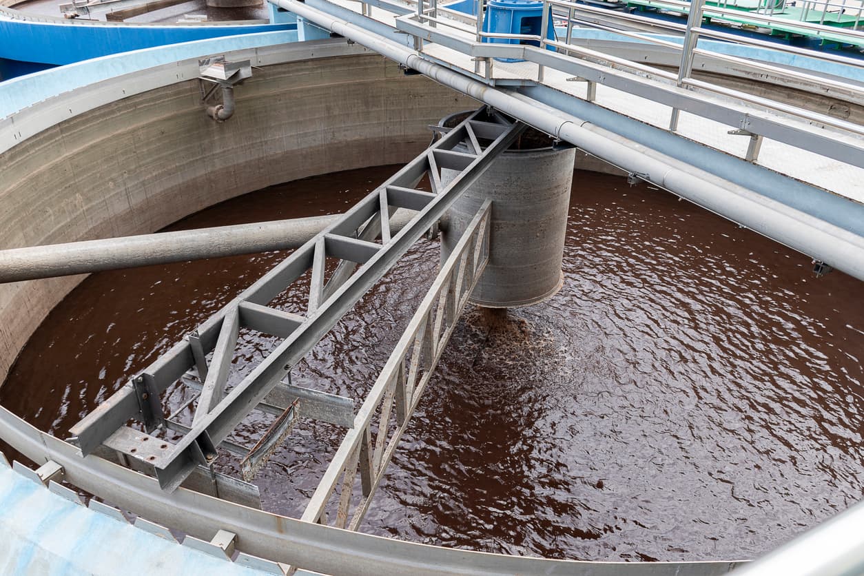 Se observa un tanque circular con agua de color marrón, en el centro hay una estructura cilíndrica gris con una base amplia. Desde la parte superior, varias estructuras metálicas se extienden hacia el borde del tanque, algunas son Tubería Helicoidal que transportan el agua. El ambiente parece ser parte de una planta de tratamiento de aguas.