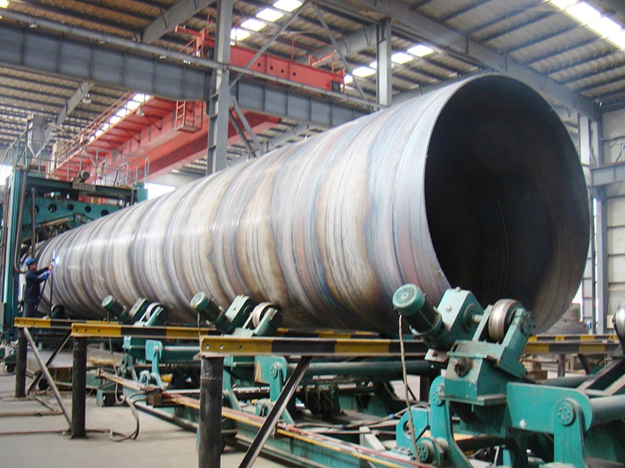 En un taller industrial, se observa una gran tubería helicoidal en proceso de fabricación. La tubería está montada sobre una estructura con rodillos, y un operario la está manipulando con herramientas. El ambiente es amplio, con techos altos y luces artificiales.