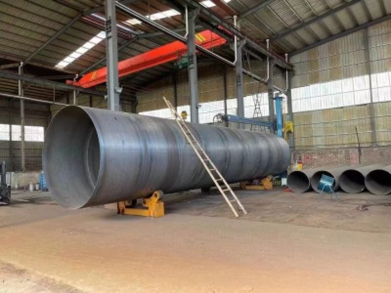En un taller industrial, hay una tubería helicoidal de gran tamaño apoyada en soportes, con una escalera de mano a su lado. Al fondo, se observan varios tubos helicoidales más pequeños alineados en el suelo. La iluminación proviene de las ventanas del establecimiento.