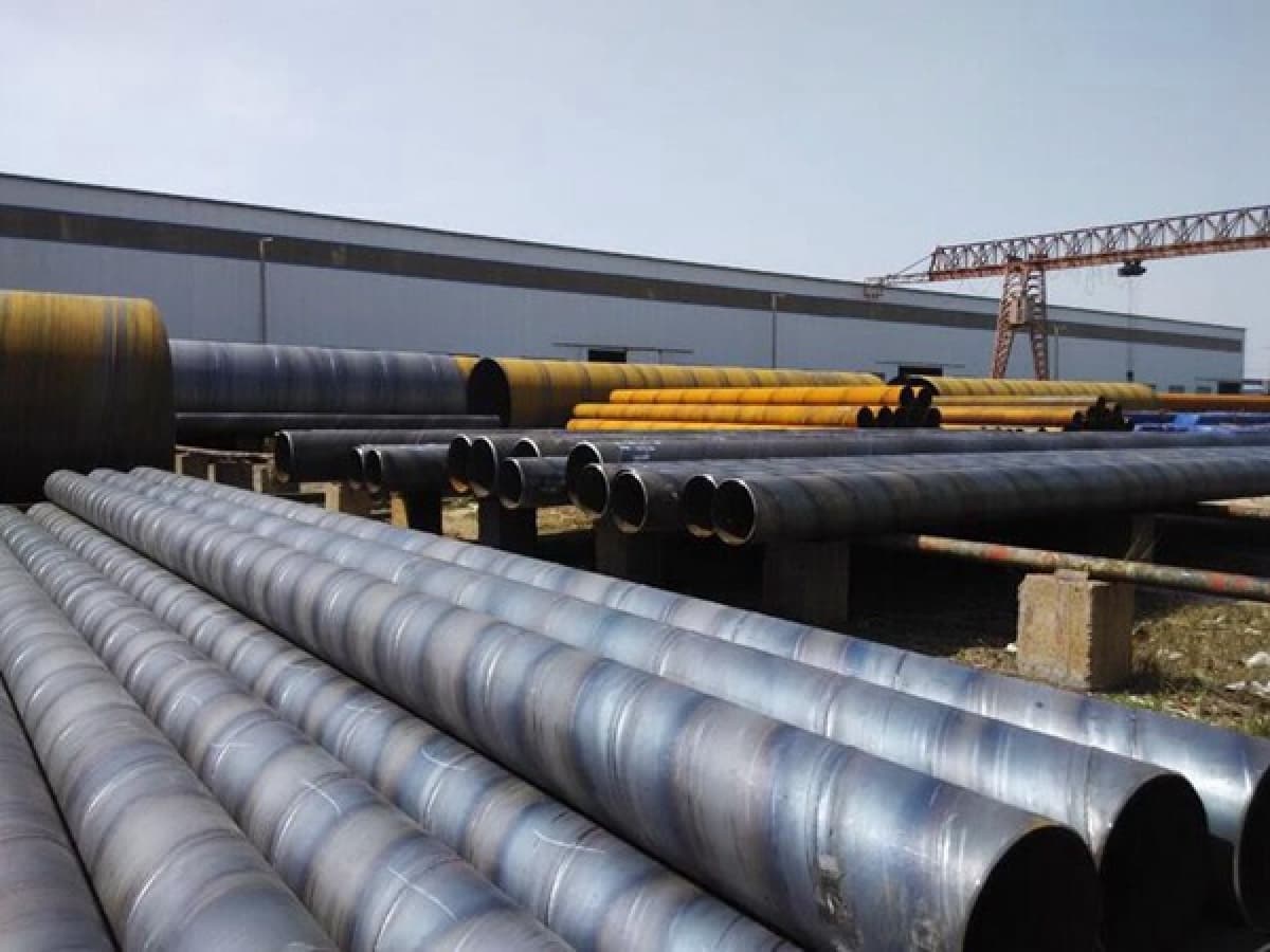 En un entorno industrial, se observan varios tubos helicoidales grandes y metálicos, apilados sobre una superficie amarilla y negra. En el fondo, hay una estructura verde que parece ser un sistema de grúas o poleas, y el ambiente es amplio, con iluminación artificial.
