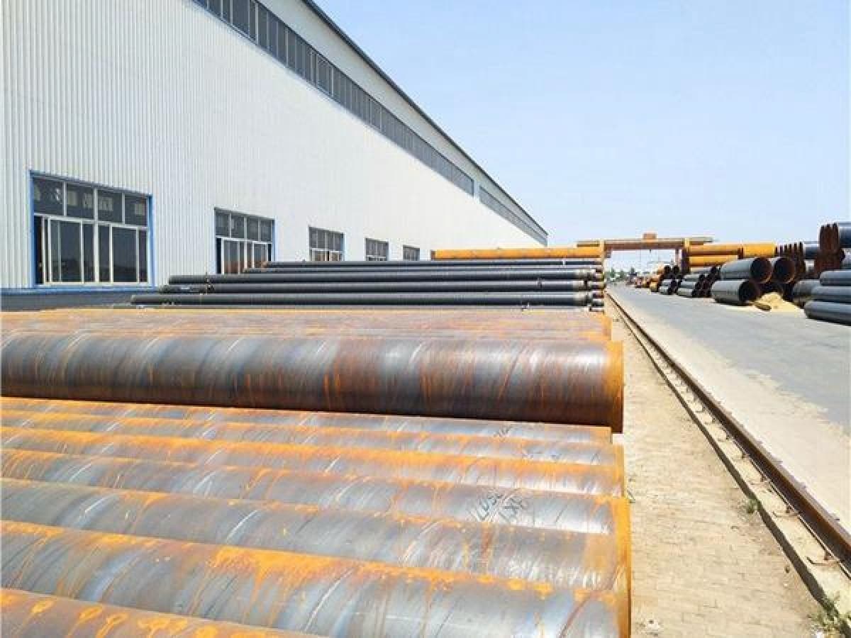 Se observan numerosos tubos helicoidales apilados en un área de trabajo al aire libre, junto a un edificio industrial. El ambiente es despejado y soleado, con un camino que se extiende entre los tubos y lleva hacia una estructura en el fondo.
