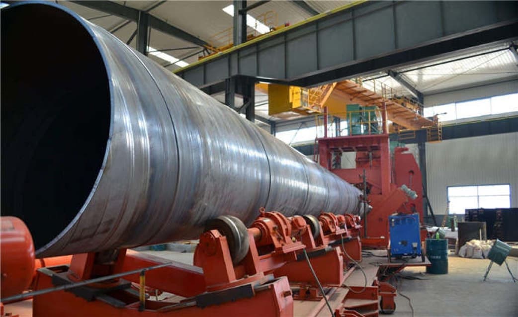 Se observa una larga tubería helicoidal en un taller industrial, apoyada sobre un soporte rojo. El ambiente es luminoso, con un techo elevado y estructuras metálicas. Al fondo, hay maquinaria y herramientas visibles, junto con algunos barriles y materiales de trabajo.