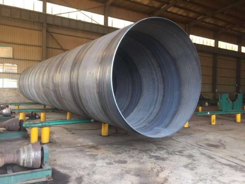 Se observa una gran tubería helicoidal de metal, ubicada en un espacio industrial. La tubería es de un diámetro considerable y está apoyada sobre soportes amarillos. El entorno es un taller con suelo de cemento y paredes de acero.