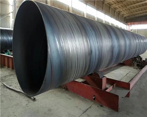 Se observa una tubería helicoidal de gran diámetro, con un acabado metálico oscuro y superficies lisas, ubicada sobre una estructura de soporte. El ambiente es industrial, con iluminación natural y un techo elevado.