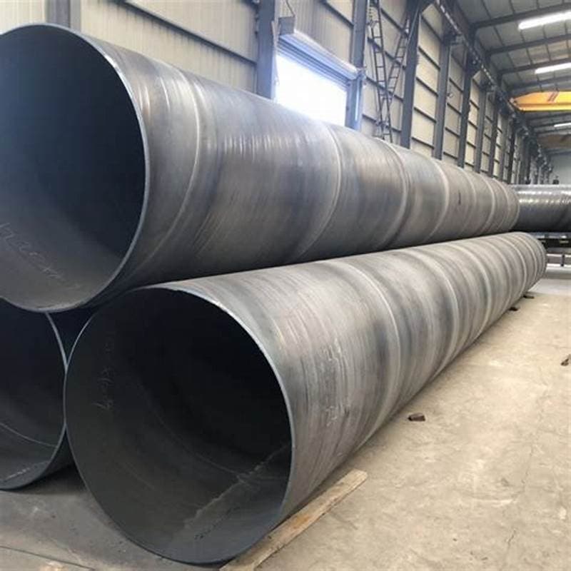 Varios tubos helicoidales de gran diámetro apilados en un espacio industrial. Las paredes de los tubos son oscuras y tienen un acabado rugoso. El entorno es un taller con iluminación natural y estructuras metálicas visibles.