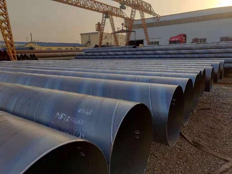 Se observan varios tubos heicoidales alineados en un área de trabajo, con grúas en la parte superior y un edificio al fondo. La iluminación es tenue, sugiriendo que es al atardecer.