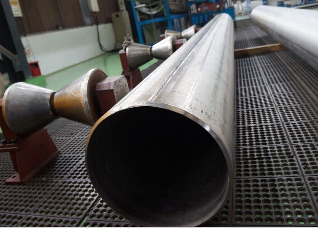 En la imagen se observan varios tubos helicoidales en primer plano, con un fondo que muestra un ambiente industrial. También hay unos accesorios con forma de embudo colocados en un soporte. El suelo está hecho de un material con perforaciones.