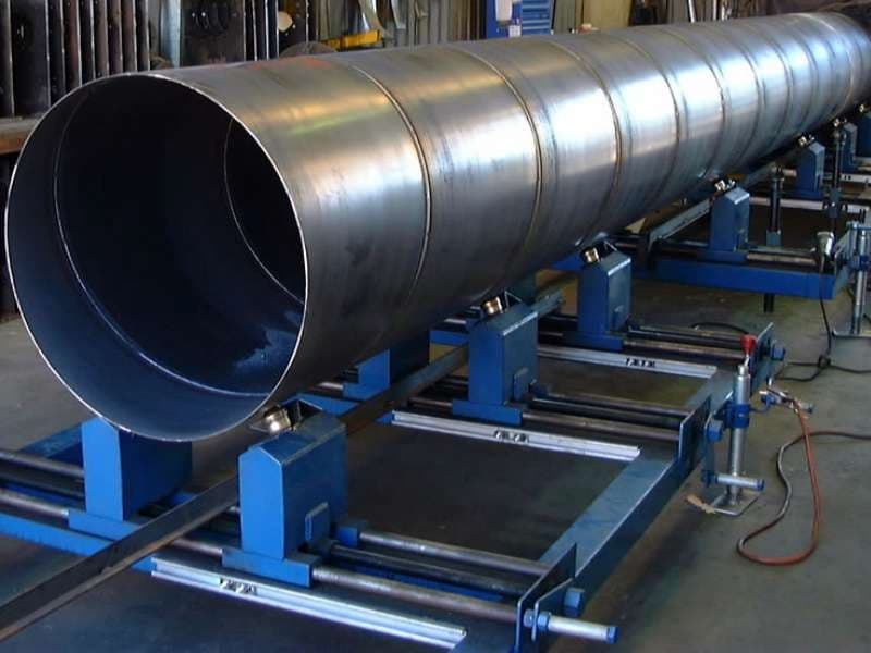 Se observa una tubería helicoidal de gran diámetro apoyada sobre una estructura azul que facilita su manipulación. La superficie de la tubería es metálica y reflectante, y el entorno parece ser un taller industrial.