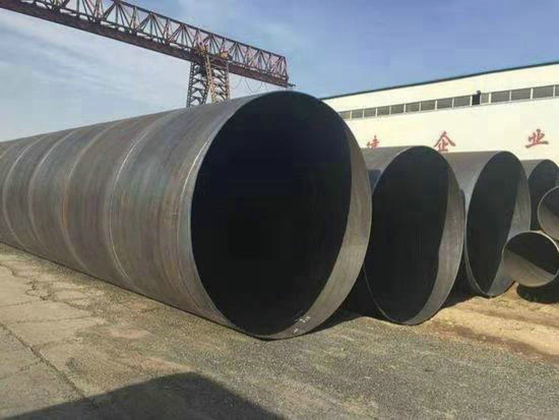 Se observan varios tubos helicoidales de gran tamaño apilados en un área de construcción, con un fondo que muestra un edificio industrial y una grúa en la parte superior. El cielo es de un color claro, lo que sugiere una buena visibilidad.