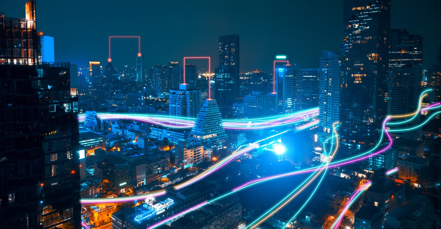 Una vista nocturna de una ciudad iluminada, con edificios altos y modernos. Se aprecian luces de colores vibrantes que parecen moverse a lo largo de las calles, creando un efecto dinámico. Algunas áreas están resaltadas con rectángulos luminosos, añadiendo un toque futurista al paisaje urbano.