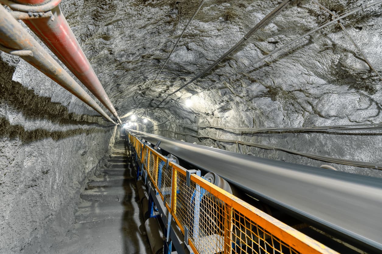 Vista de un túnel subterráneo con paredes rugosas y oscuras. A la izquierda se observan tubos heicoidales rojos, mientras que en el centro hay una cinta transportadora en movimiento protegida por una barandilla de malla metálica. Se aprecian luces en el túnel que iluminan el camino.