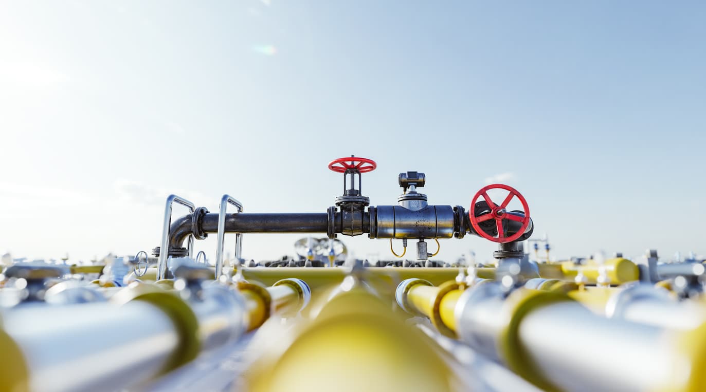 Una estructura metálica con válvulas rojas se encuentra en el centro, rodeada de tubos heicoidales en el primer plano. Al fondo, se puede ver un paisaje industrial bajo un cielo despejado.