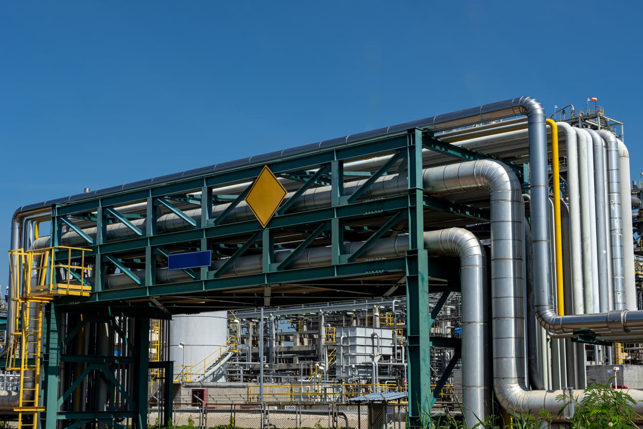 Estructura metálica soporte con tubos helicoidales conectados, señales de advertencia en color amarillo y azul, y un fondo industrial con más instalaciones y tuberías. El cielo es despejado y azul.